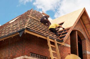 Roofing worker rolls synthetic underlayment across a residential roof deck beside stacked shingles.