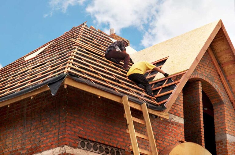 Roofing worker rolls synthetic underlayment across a residential roof deck beside stacked shingles.